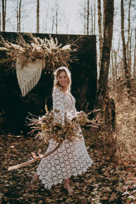 laughing blonde boho bride in crocheted dress in the fall in the woods on a sunny day with a foraged dried flower bouquet with fall colored trailing ribbons
