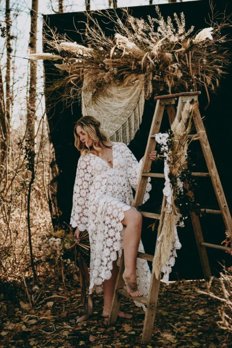 fall boho bride in the woods with rustic wooden ladder wearing a crocheted dress with a dried floral backdrop with crochet shawl accents