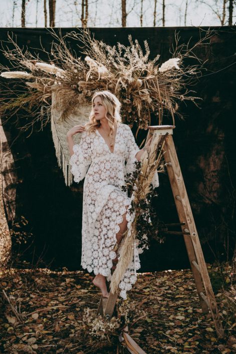 blonde boho bride on ladder with dried pampas grass and dried floral backdrop in the fall in the woods