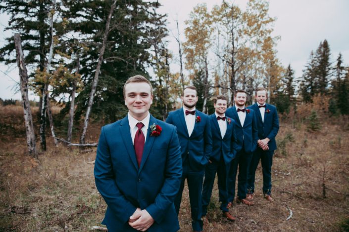 groom and groomsmen in navy suits with red bow ties and red spray rose boutonniere in line on a fall day in a forest