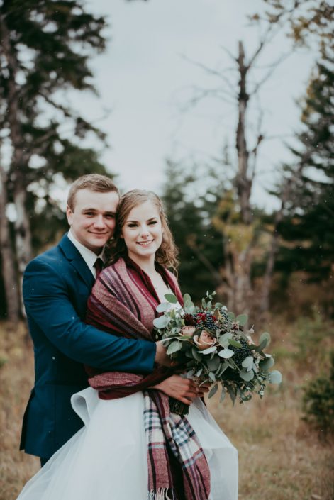 bride and groom photo cheek to cheek with plaid shawl and groom wearing a blue suit bridal bouquet with mixed eucalyptus greenery and red roses and burgundy dahlias and amnesia roses against a natural backdrop in Alberta
