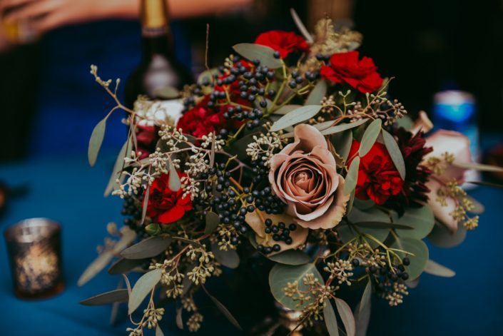 close up of low centerpiece with navy viburnum berry red carnations burgundy dahlias amnesia roses and mixed eucalyptus greenery in a compote vase on a blue tablecloth