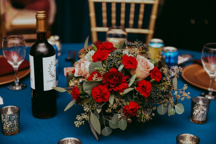 Low Centerpiece with red carnations quicksand roses and viburnum berries and mixed eucalyptus greenery on blue tablecloth with gold chairs close up shot