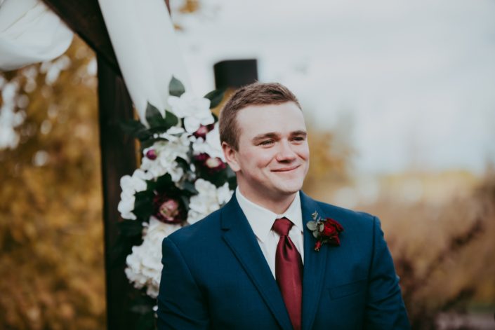 portrait of groom in navy suit with red spray rose boutonniere and red tie smiling in front of rustic archway