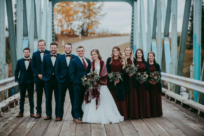 bridal party on bridge with blue suits and bowties and burgandy bridesmaid dresses and shawls and bride with plaid shawl and bouquets with mixed eucalyptus and red roses and burgundy dahlias an blue viburnum berries