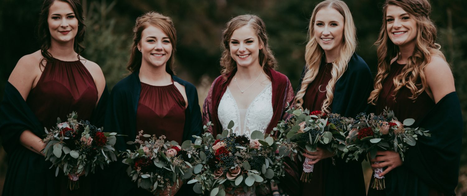 bridal party picture with burgundy bridesmaid dresses and green shawls and bride with plaid shawl and bouquets with red roses burgundy dahlias navy viburnum berries and mixed eucalyptus greenery against forest backdrop in alberta