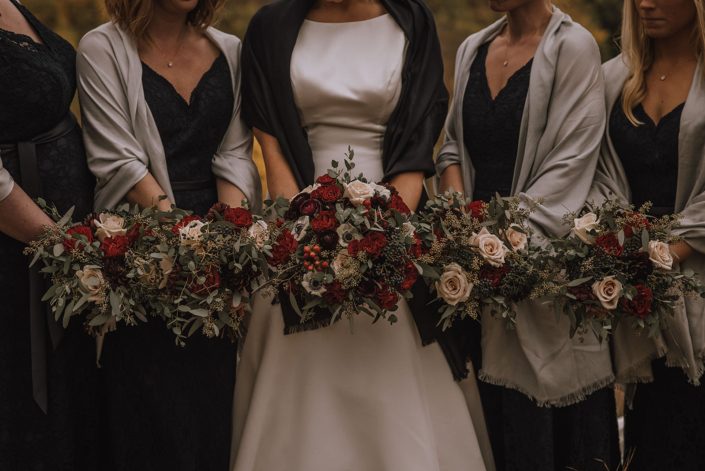 autumn bridal party in black dresses with bride and bridesmaid bouquets designed with ivory sahara roses red hearts garden roses and deep burgundy ranunculus ad accented with eucalyptus greenery