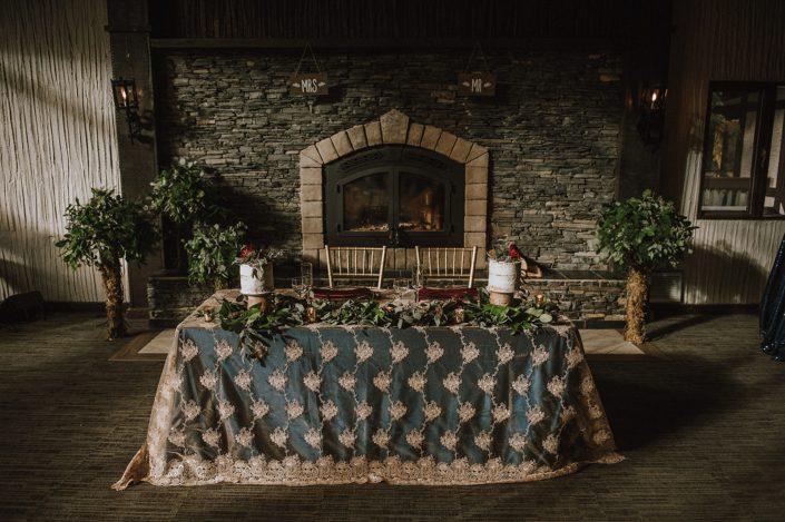 Sweetheart table at Canyon Ski Resort in Red Deer in front of fireplace with mixed greenery trees and greenery garland and his and hers naked cakes