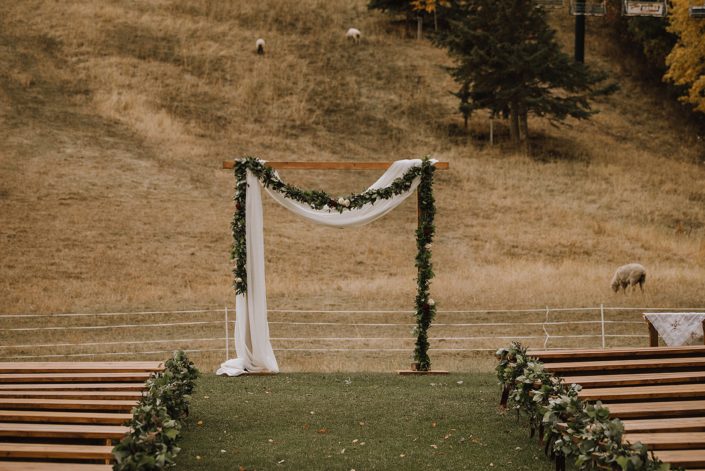 Cermeony site at CAnyon Ski resort decorated with frsh greenery garland and voile draping over the rustic wooden archway with sheep in the background