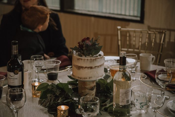 wedding cake centerpiece creathed with a naked cake on a birch log with a ring of eucalyptus greenery below and accented by a burgundy hearts garden rose and navy eryngium bloom