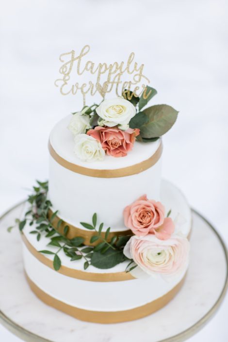 winter wedding cake with white fondant and gold trim accented by gunni eucalyptus, peach spray roses and ivory hanoi ranunculus