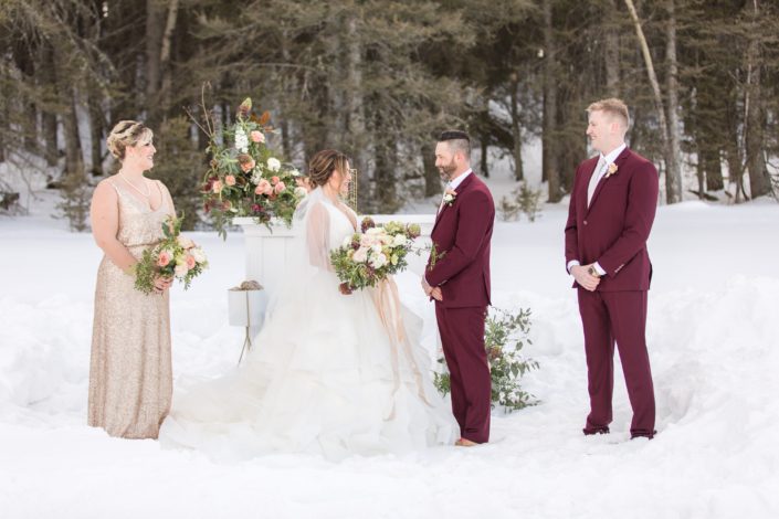 Winter wedding party with bridesmaids in blush sequin and groomsmen in burgundy suits holding flowers in brugundy, blush and ivory in front of a white mantle