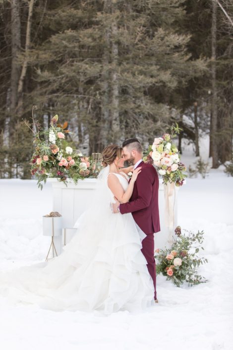 Bride and groom in winter wedding styled shoot in front of white mantle with flower arrangements in peach, blush and ivory