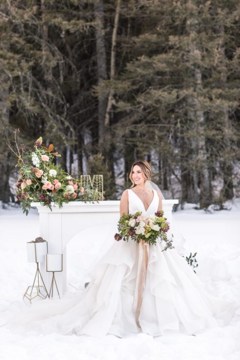 Bride in winter wedding styled shoot holding bouquet in front of mantle with floral arrangements and bridal bouquet in burgundy, blush and white