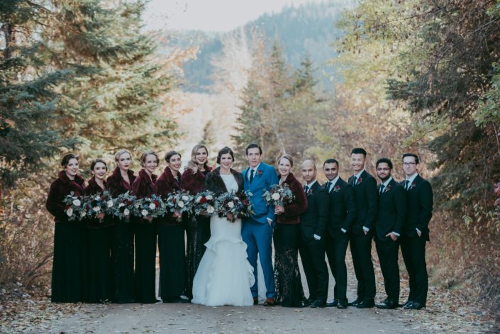 very large bridal party photo with7 bridesmaids groom in blue suit with a red rose boutonniere groomsmen in black suits with hands in pockets bridesmaids in black sequin dresses and fur coats fall wedding on road with tree lined sides