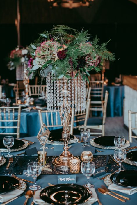 Glamorous tall centerpiece with gold and crystal accents with red roses hydrangea and mixed greenery blue table cloths and black chargers and a black sequin runner canyon ski resort wedding in red deer, Alberta