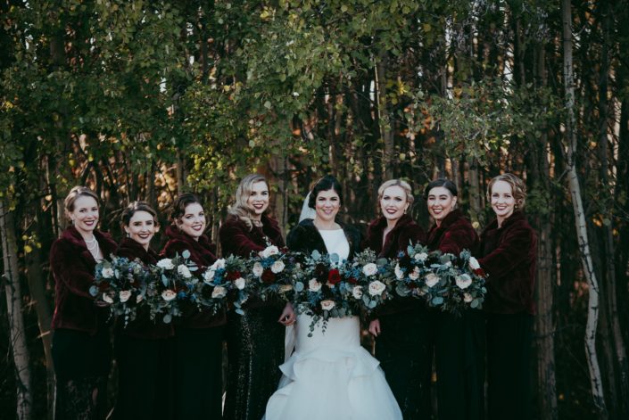 bride and bridesmaids photo with fur coats against a treed background smiling holding out matching bouquets with mixed eucalyptus greenery and red and blush roses large bridal party