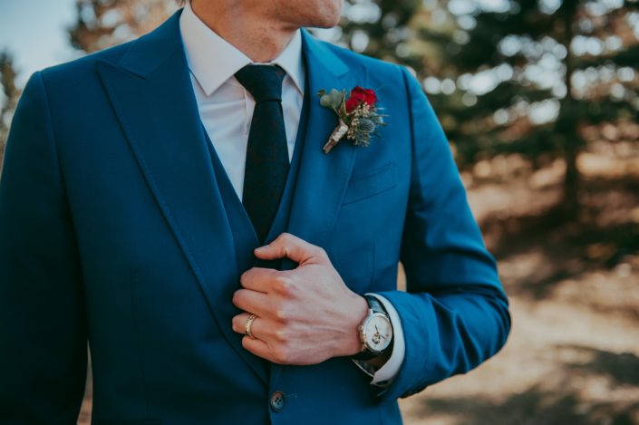 Groom in blue suit wearing a red rose and eryngium boutonniere and watch