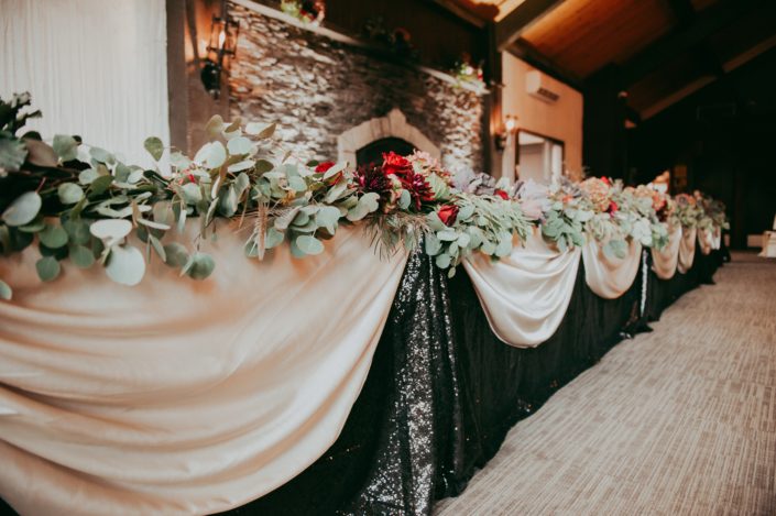 Head Tab;e draped with champagne satin and black sequin linens and a fresh mixed eucalyptus greenery garland accented by burgundy roses and dahlia at Canyon Ski Resort in Red Deer