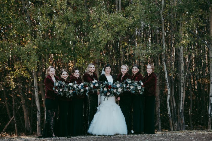 Bride and bridesmaid photo with fur coats and black sequin dresses against a forest backdrop bouquets with red roses, quicksand roses, eryngium and mixed eucalyptus greenery fall wedding