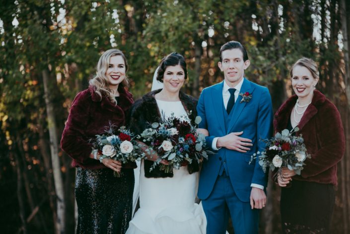 Fall bridal party picture with furn jackets and black sequin dresses groom in blue suit with red spray rose boutonniere bouquets with red roses, quicksand roses, burgundy dahlias and mixed eucalyptus