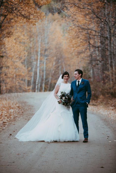 Bride and Groom picture in fall on a road lined with fall trees holding a wedding bouquet with red roses and blue eryngium groom wearing blue suit an brown shoes at Canyon Ski Resort in Red Deer, Alberta