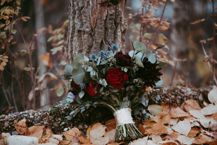 Handtied wedding bouquet in blue and burgandy with red garden roses burgundy dahlias eryngium mixed eucalyptus in rustic setting displayed in a forest in fall fall wedding at canyon ski resort in red deer, alberta