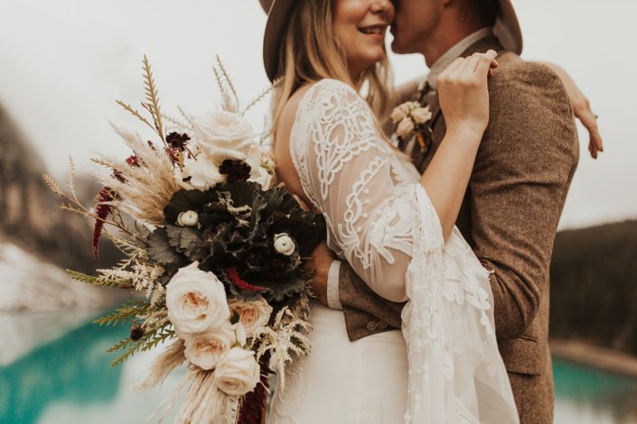 Moraine Lake Elopement Styled Shoot - male model wearing a hat and an ivory boutonniere, brown jacket and bolo tie. Girl wearing a hat and an ivory lace bridal gown while holding pampas grass bouquet with blush roses and pops of red flowers