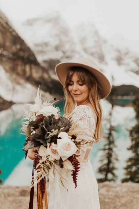 Moraine Lake Elopement Styled Shoot - model wearing ivory bridal gown and hat holding pampas grass bouquet with blush roses and pops of red tied with trailing ribbons.