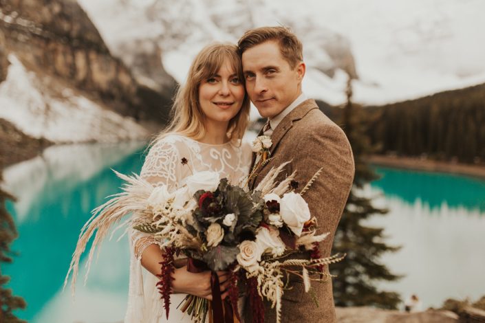 Moraine Lake Elopement Styled Shoot - male model wearing ivory boutonniere, brown jacket and bolo tie. Girl wearing ivory lace bridal gown while holding pampas grass bouquet with blush roses and pops of red flowers