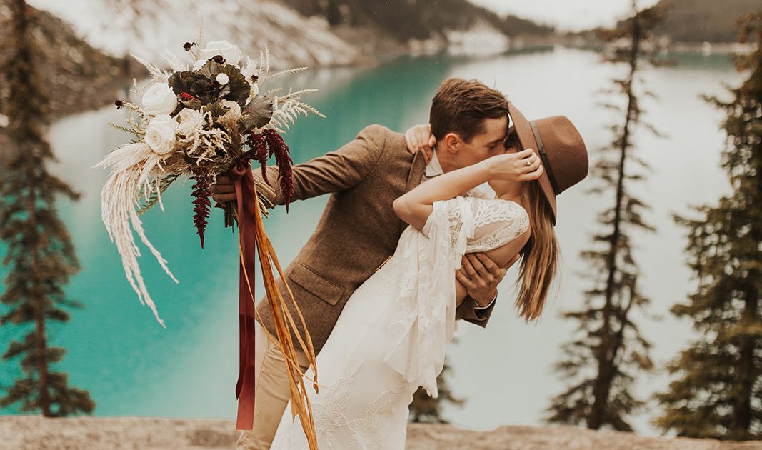 Moraine Lake Elopement Styled Shoot - couple kissing and girl wearing ivory bridal gown and man holding pampas grass bouquet with blush roses and pops of red tied with trailing ribbons.