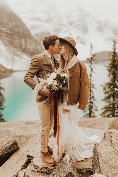 Moraine Lake Elopement Styled Shoot - couple kissing and girl wearing brown jacket and ivory bridal gown and hat holding pampas grass bouquet with blush roses and pops of red tied with trailing ribbons.