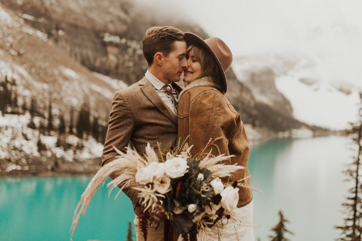 Moraine Lake Elopement Styled Shoot - couple embracing and girl wearing brown jacket and ivory bridal gown and hat holding pampas grass bouquet with blush roses and pops of red tied with trailing ribbons.