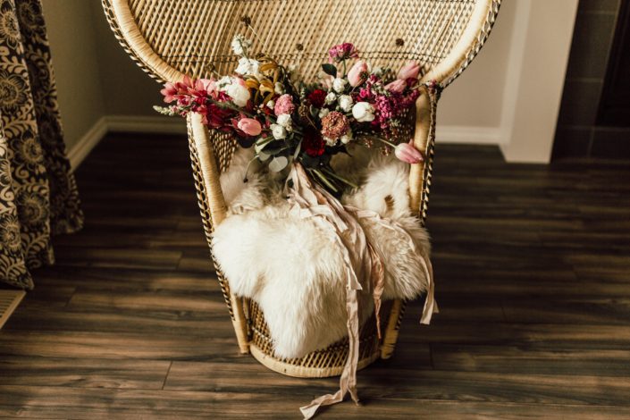 Red and Pink Styled Shoot - bouquet made of pink and white tulips, scabiosa, roses, and greenery tied with trailing ribbon. It is atop a vintage boho rattan chair covered in a sheepskin rug.