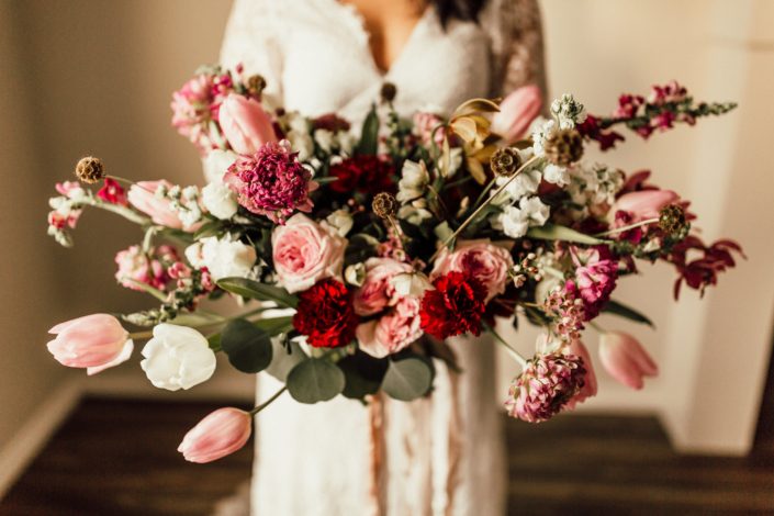Red and Pink Styled Shoot - Model wearing an ivory lace bridal gown and holding a bouquet made of pink and white tulips, scabiosa, roses, and greenery tied with trailing ribbon.
