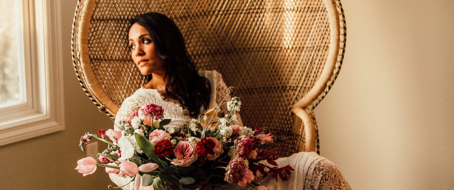 Red and Pink Styled Shoot - Model sitting in vintage rattan chair wearing ivory lace bridal gown and holding a bouquet made of pink and white tulips, scabiosa, roses, and greenery tied with trailing ribbon.