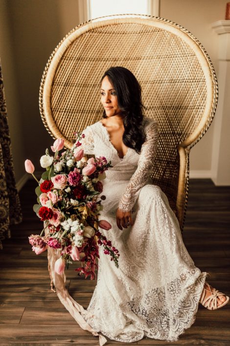 Red and Pink Styled Shoot - Model sitting in vintage rattan chair wearing ivory lace bridal gown and holding a bouquet made of pink and white tulips, scabiosa, roses, and greenery tied with trailing ribbon.