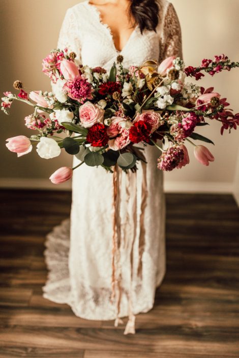 Red and Pink Styled Shoot - Model wearing an ivory lace bridal gown and holding a bouquet made of pink and white tulips, scabiosa, roses, and greenery tied with trailing ribbon.