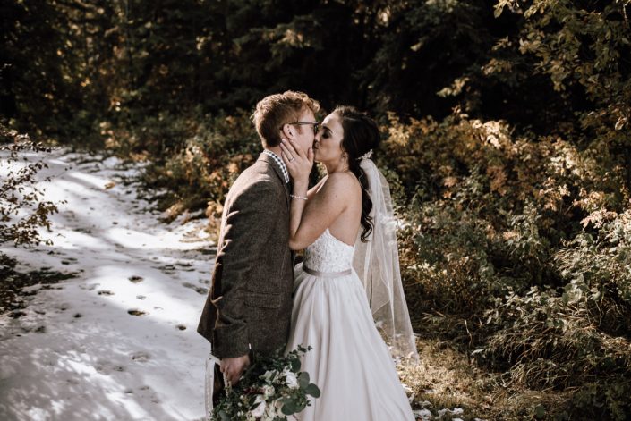 bride kissing groom in the winter with snow holding a white and greenery bouquet