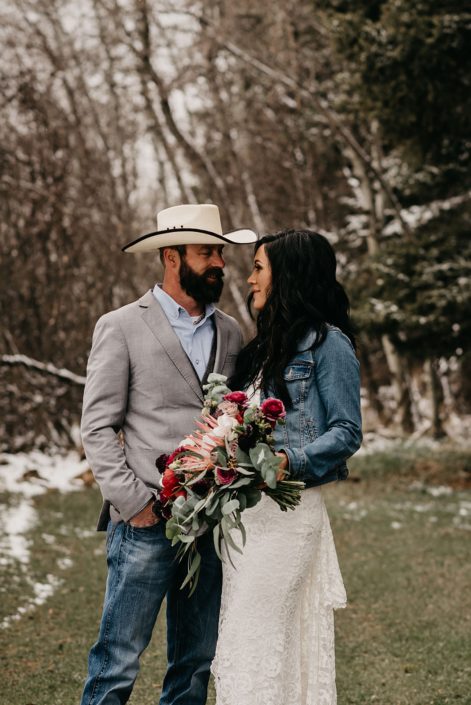 Rustic Boho Chic Wedding - Bride and groom standing together. Groom wearing cowboy hat, sport coat and jeans. Bride wearing ivory lace dress and jean jacket holding bridal bouquet made of king protea, red peonies, pink roses, plum scabiosa, panda anenome, and eucalyptus greenery.