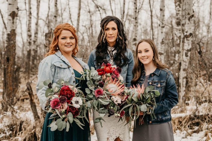 Rustic Boho Chic Wedding - Bride holding bridal bouquet made of king protea, red peonies, pink roses, plum scabiosa, panda anenome, and eucalyptus greenery. She is standing next to her bridesmaid who is holding a bouquet made of red peony, blush and pink roses, and eucalyptus greenery. On her other side is her flower girl who is holding a bouquet made of single king protea and eucalyptus greenery. The bride, bridesmaid and flower girl are all wearing jean jackets.