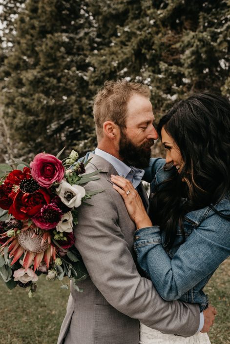 Rustic Boho Chic Wedding - Bride and groom standing together. Bride wearing a jean jacket holding bridal bouquet made of king protea, red peonies, pink roses, plum scabiosa, panda anenome, and eucalyptus greenery.
