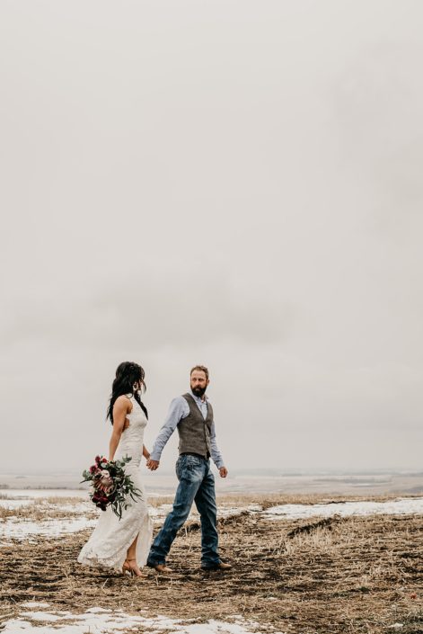 Rustic Boho Chic Wedding - Bride and groom walking together in a snow covered field. Groom wearing vest and jeans. Bride wearing ivory lace dress holding bridal bouquet made of king protea, red peonies, pink roses, plum scabiosa, panda anenome, and eucalyptus greenery.
