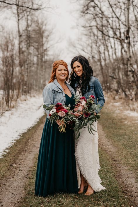 Rustic Boho Chic Wedding - Bride wearing ivory lace dress and jean jacket holding bridal bouquet made of king protea, red peonies, pink roses, plum scabiosa, panda anenome, and eucalyptus greenery standing next to her bridesmaid who is holding a bouquet made of red peony, blush and pink roses, and eucalyptus greenery.