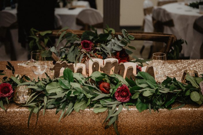 Rustic Boho Chic Wedding - Mr and Mrs sign with a long garland made of fresh greenery and red flowers on a gold sequin covered head table.