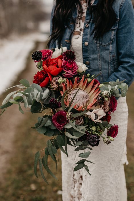Rustic Boho Chic Wedding - Bride wearing ivory lace dress and jean jacket holding bridal bouquet made of king protea, red peonies, pink roses, plum scabiosa, panda anenome, and eucalyptus greenery.