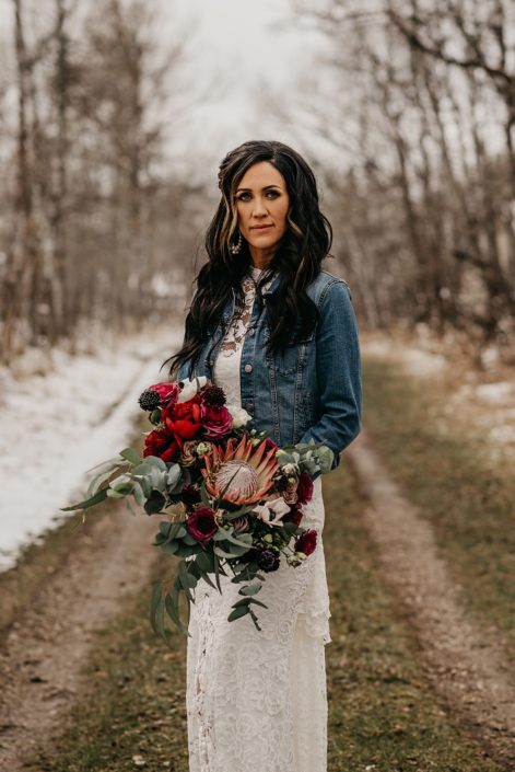 Rustic Boho Chic Wedding - Bride wearing ivory lace dress and jean jacket holding bridal bouquet made of king protea, red peonies, pink roses, plum scabiosa, panda anenome, and eucalyptus greenery.