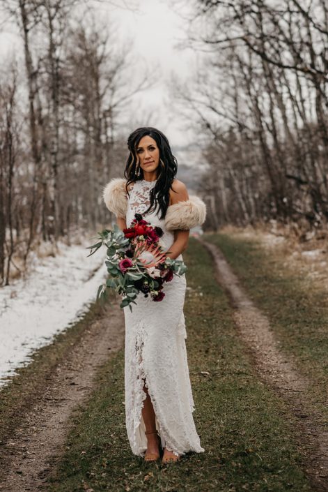 Rustic Boho Chic Wedding - Bride wearing ivory lace dress and fur shawl covering her shoulders while holding bridal bouquet made of king protea, red peonies, pink roses, plum scabiosa, panda anenome, and eucalyptus greenery.