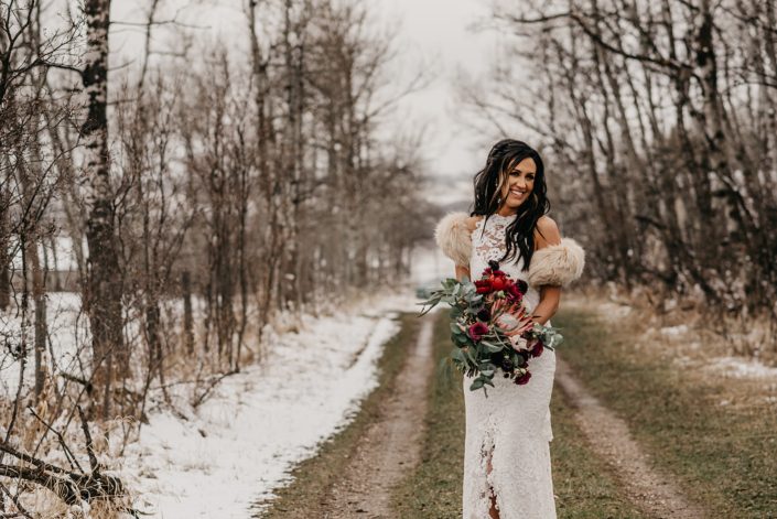 Rustic Boho Chic Wedding - Bride wearing ivory lace dress and fur shawl covering her shoulders while holding bridal bouquet made of king protea, red peonies, pink roses, plum scabiosa, panda anenome, and eucalyptus greenery.