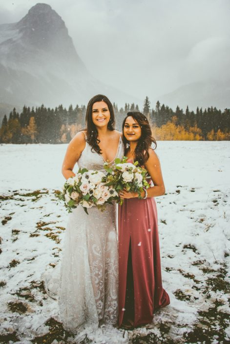 Bride and bridesmaid of the Blush and Mauve Canmore Wedding standing in the snow in the Rocky Mountains. They each are holding a fresh hand-tied bouquet made with blush, white and mauve flowers and greenery.